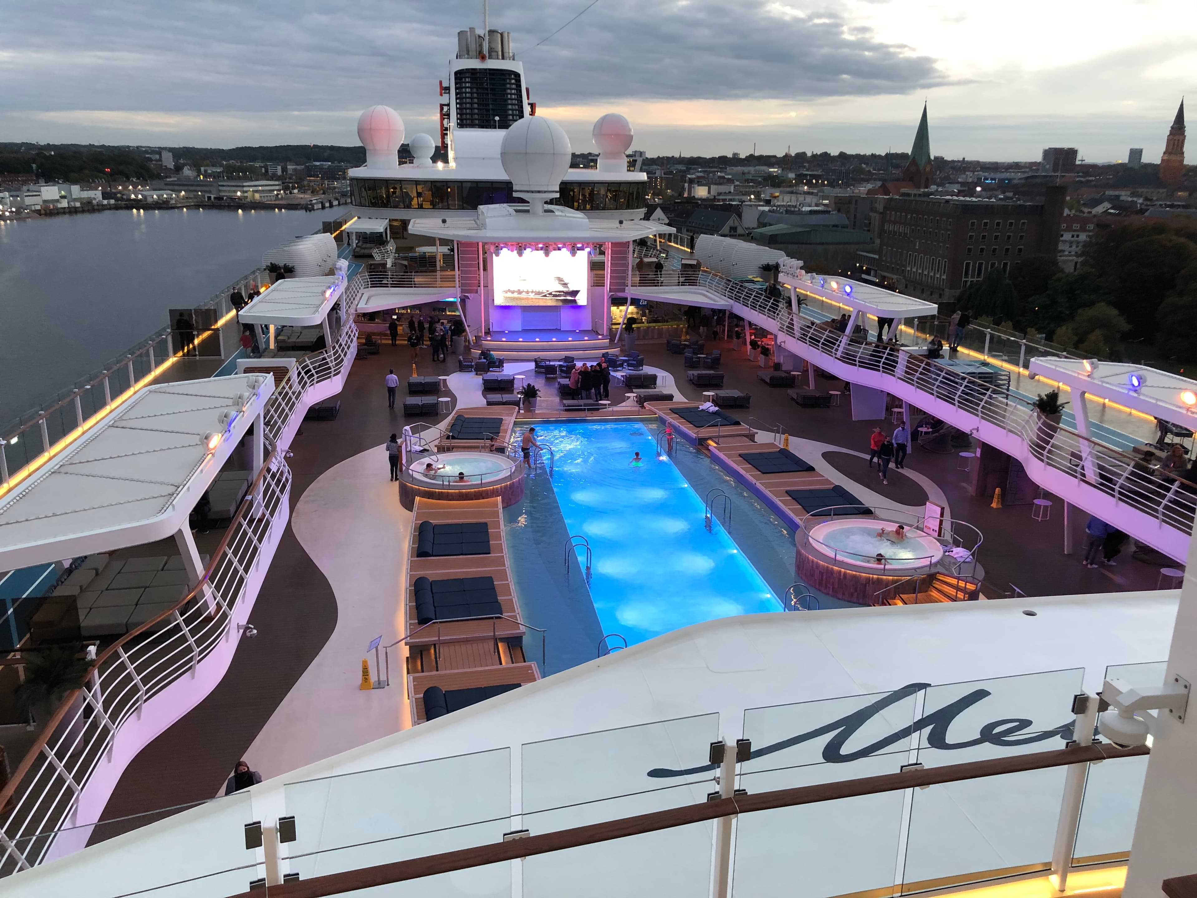 Mein Schiff Pooldeck von oben im Abenddämmerlicht — beleuchteter Pool, Whirlpools und Bühne mit Kiel im Hintergrund